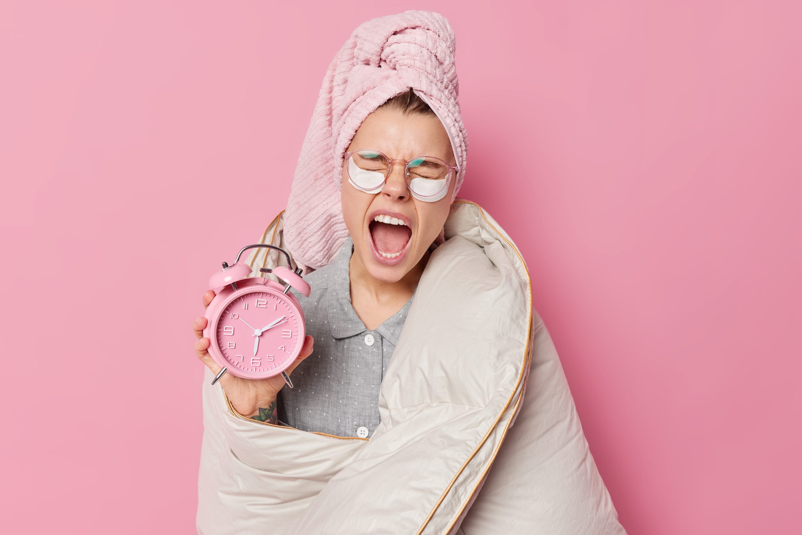early awakening concept sleepy young european woman yawns keeps mouth widely opened holds alarm clock wrapped blanket wears bath towel head after taking shower isolated pink background scaled